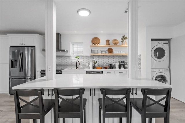 kitchen with light wood finished floors, wall chimney range hood, stainless steel fridge with ice dispenser, decorative backsplash, and stacked washing maching and dryer