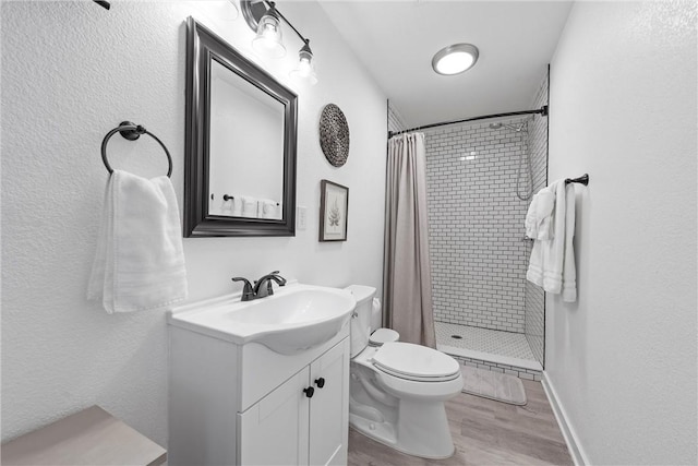 bathroom with toilet, wood finished floors, a tile shower, baseboards, and vanity