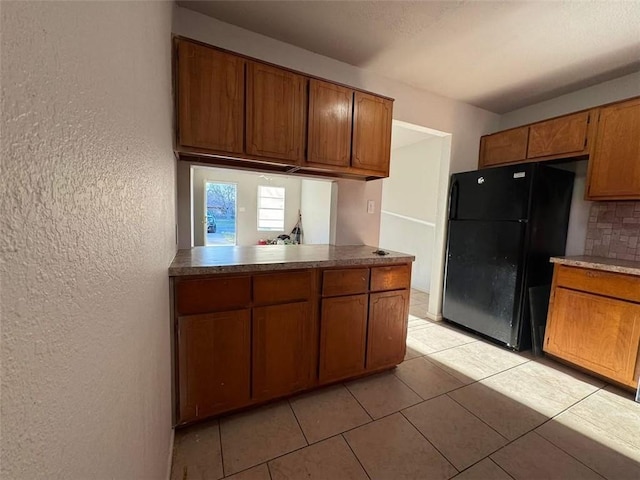 kitchen featuring backsplash, a textured wall, freestanding refrigerator, light tile patterned flooring, and brown cabinetry