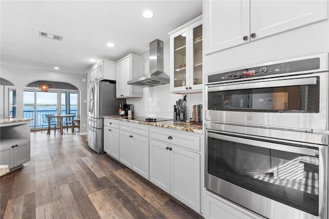 kitchen with light stone countertops, stainless steel appliances, wall chimney range hood, white cabinets, and a water view