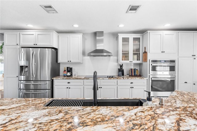 kitchen with appliances with stainless steel finishes, white cabinetry, wall chimney exhaust hood, and light stone counters