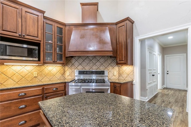 kitchen featuring premium range hood, crown molding, light hardwood / wood-style floors, dark stone counters, and appliances with stainless steel finishes