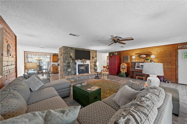 living room with ceiling fan with notable chandelier, a stone fireplace, a textured ceiling, and wooden walls