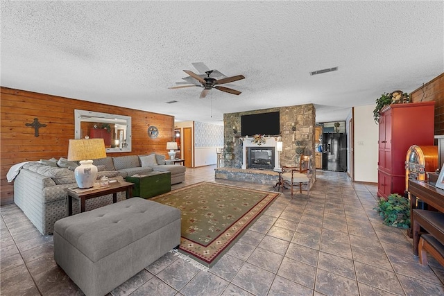 tiled living room featuring a textured ceiling, a stone fireplace, ceiling fan, and wooden walls