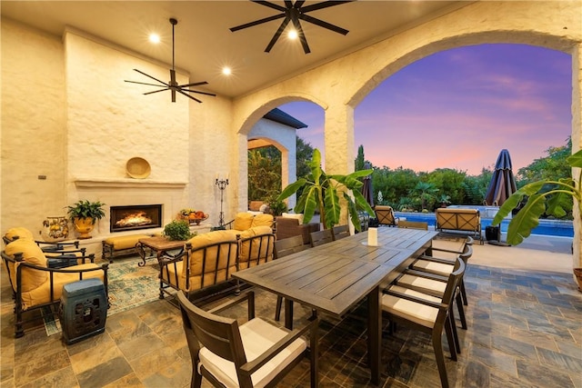 patio terrace at dusk with a jacuzzi, an outdoor living space with a fireplace, and ceiling fan