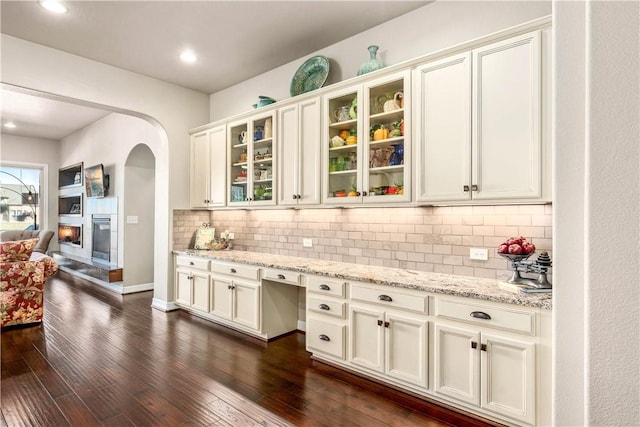 kitchen featuring tasteful backsplash, glass insert cabinets, dark wood-type flooring, light stone counters, and arched walkways