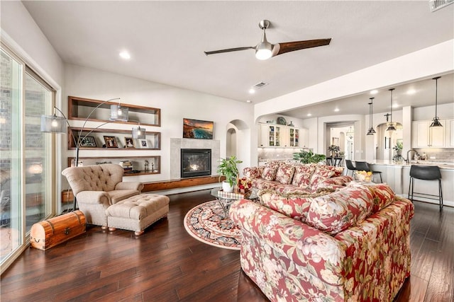 living area featuring a glass covered fireplace, dark wood-type flooring, visible vents, and ceiling fan