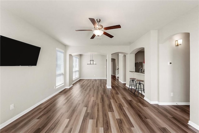 unfurnished living room with wood finished floors, ceiling fan with notable chandelier, arched walkways, and baseboards