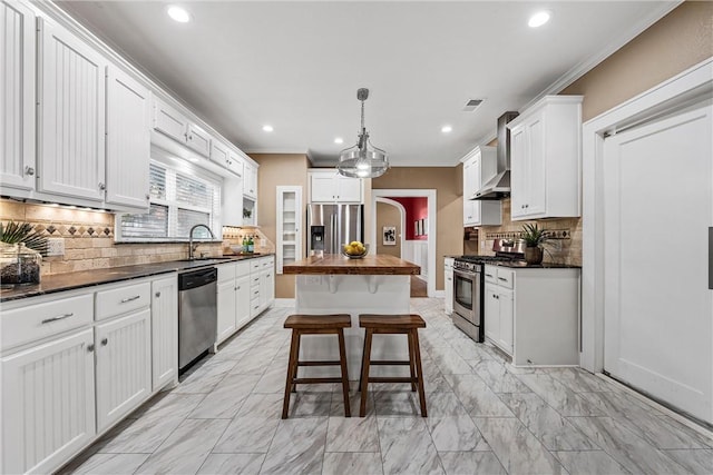 kitchen featuring wall chimney range hood, white cabinets, pendant lighting, a kitchen island, and appliances with stainless steel finishes