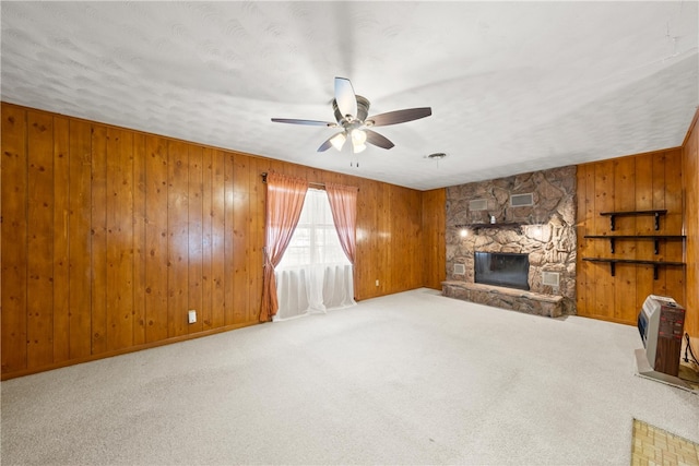 unfurnished living room featuring wooden walls, carpet floors, a stone fireplace, heating unit, and a ceiling fan