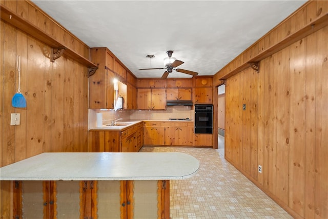 kitchen with brown cabinets, under cabinet range hood, wood walls, light countertops, and ceiling fan