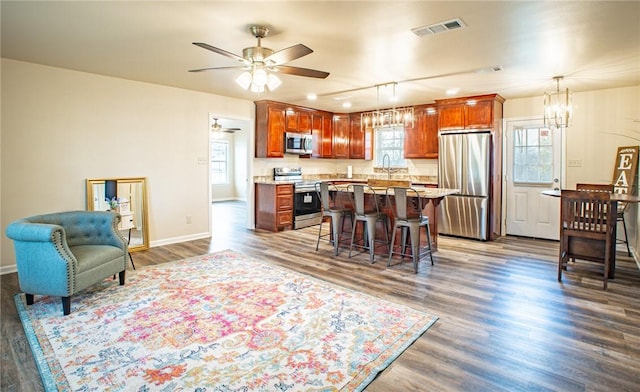 kitchen featuring dark wood-type flooring, a breakfast bar, hanging light fixtures, stainless steel appliances, and ceiling fan with notable chandelier