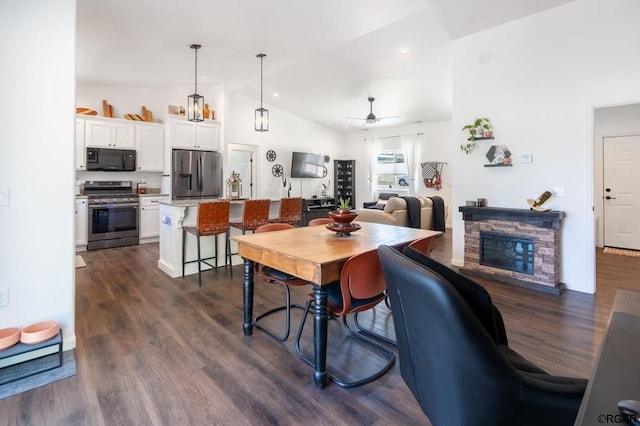 dining room featuring dark wood-type flooring, ceiling fan, a stone fireplace, and vaulted ceiling