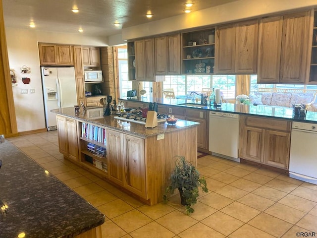 kitchen with light tile patterned floors, a center island with sink, white appliances, and dark stone counters