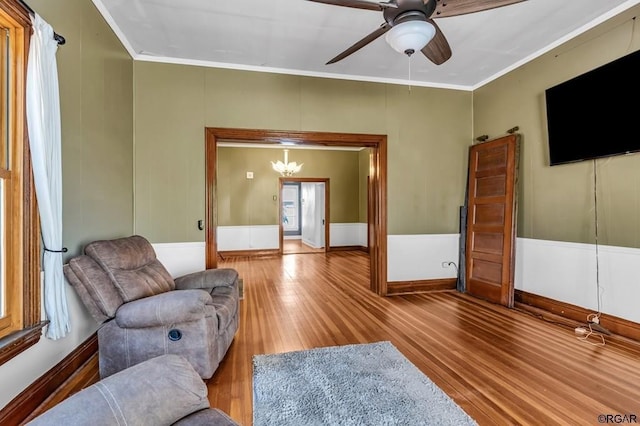 living area with ceiling fan with notable chandelier, wainscoting, crown molding, and wood finished floors