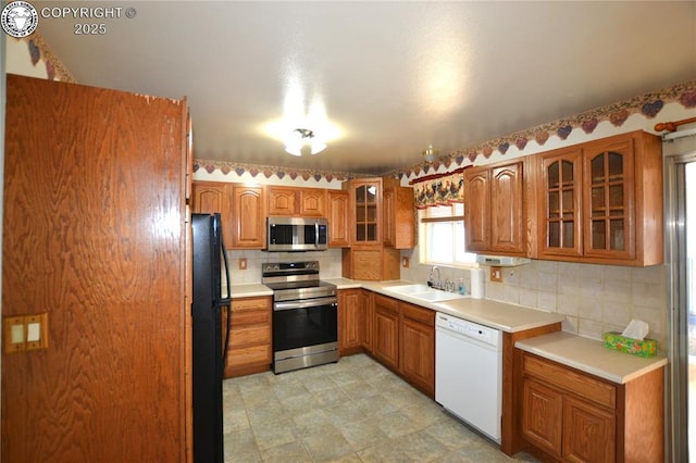 kitchen with appliances with stainless steel finishes, tasteful backsplash, brown cabinets, a sink, and glass insert cabinets