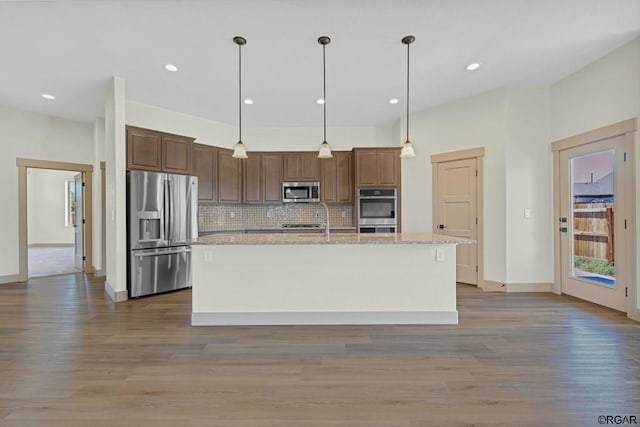 kitchen with light stone counters, tasteful backsplash, pendant lighting, stainless steel appliances, and a kitchen island with sink