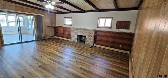 unfurnished living room with a stone fireplace, hardwood / wood-style floors, wooden walls, lofted ceiling with beams, and ceiling fan