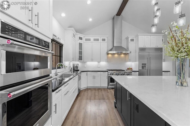 kitchen featuring premium appliances, glass insert cabinets, hanging light fixtures, wall chimney range hood, and white cabinetry