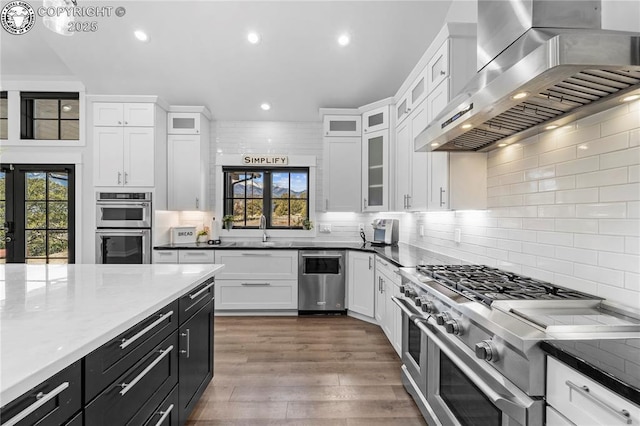 kitchen featuring range hood, stainless steel appliances, glass insert cabinets, white cabinetry, and dark cabinetry