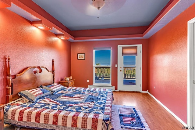 bedroom featuring light hardwood / wood-style flooring, ceiling fan, baseboard heating, access to exterior, and a raised ceiling