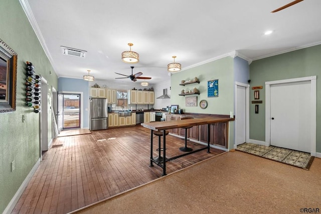 kitchen with wall chimney exhaust hood, crown molding, appliances with stainless steel finishes, ceiling fan, and cream cabinetry