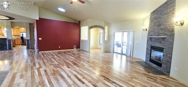 unfurnished living room with arched walkways, a stone fireplace, light wood-style flooring, a ceiling fan, and baseboards