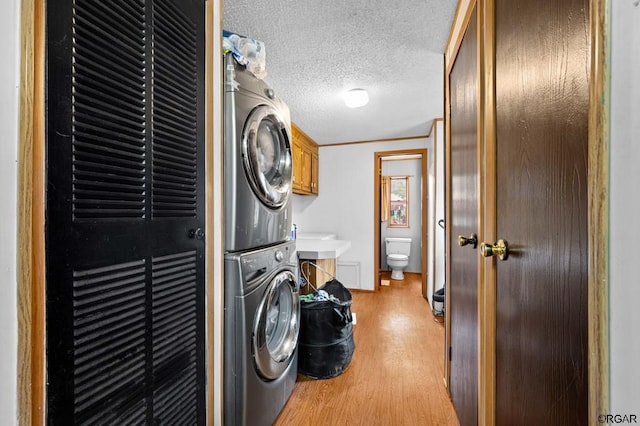 laundry room featuring stacked washer and dryer, crown molding, cabinets, a textured ceiling, and light wood-type flooring