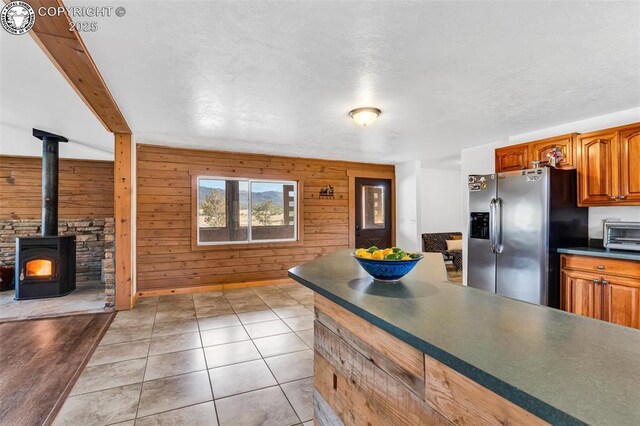 kitchen featuring stainless steel refrigerator with ice dispenser, wood walls, light tile patterned floors, a wood stove, and beam ceiling