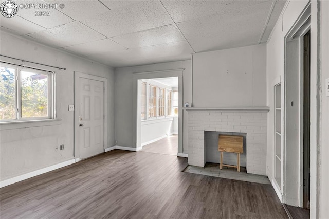 unfurnished living room with wood finished floors, baseboards, a drop ceiling, and a brick fireplace