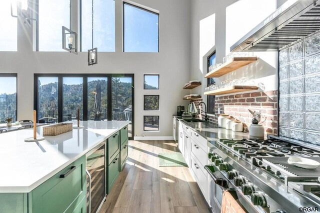 kitchen with stainless steel stove, wine cooler, green cabinets, wall chimney range hood, and light wood-type flooring