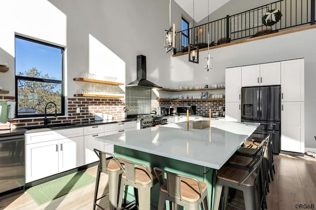 kitchen featuring white cabinetry, an island with sink, a breakfast bar area, black appliances, and wall chimney range hood