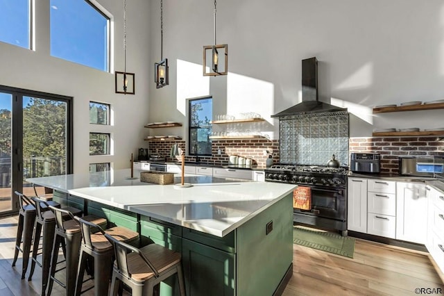 kitchen with wall chimney exhaust hood, high end range, hanging light fixtures, a kitchen island with sink, and white cabinets