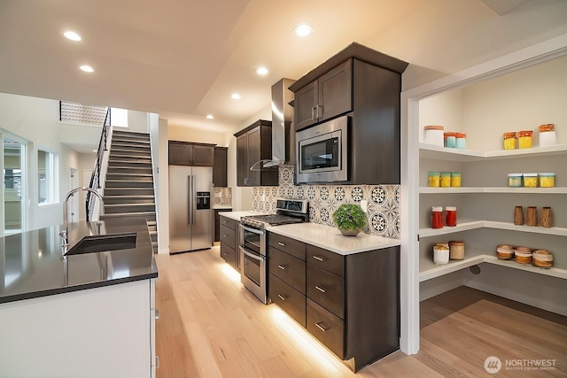 kitchen featuring appliances with stainless steel finishes, dark brown cabinetry, a sink, light wood-type flooring, and wall chimney exhaust hood