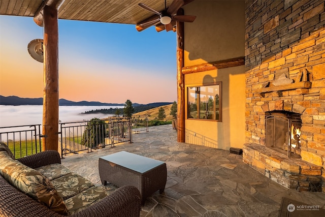 patio terrace at dusk with a mountain view, an outdoor stone fireplace, and ceiling fan