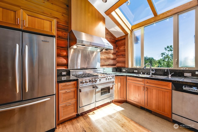 kitchen featuring stainless steel appliances, vaulted ceiling with skylight, light wood-type flooring, wall chimney range hood, and rustic walls