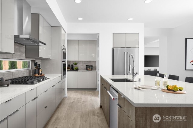 kitchen with a kitchen island with sink, appliances with stainless steel finishes, wall chimney exhaust hood, light hardwood / wood-style flooring, and tasteful backsplash