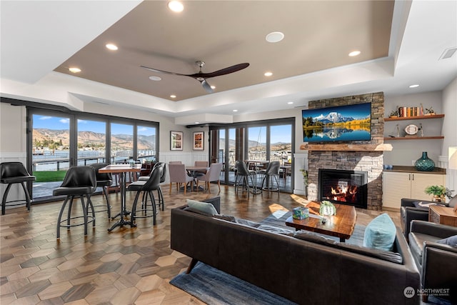 tiled living room with a raised ceiling, plenty of natural light, ceiling fan, and a stone fireplace