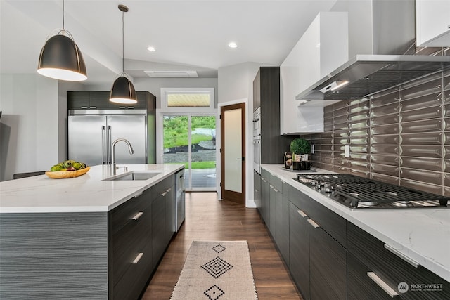 kitchen featuring wall chimney exhaust hood, stainless steel appliances, sink, pendant lighting, and a center island with sink