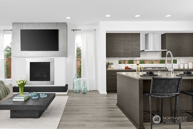 kitchen with dark brown cabinetry, a breakfast bar area, light wood-type flooring, light stone countertops, and wall chimney exhaust hood