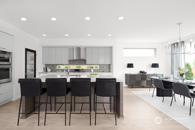 kitchen featuring stainless steel double oven, gray cabinetry, a breakfast bar, light hardwood / wood-style floors, and wall chimney range hood