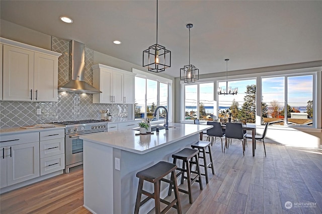 kitchen with stainless steel range, wall chimney exhaust hood, light hardwood / wood-style floors, a center island with sink, and white cabinets