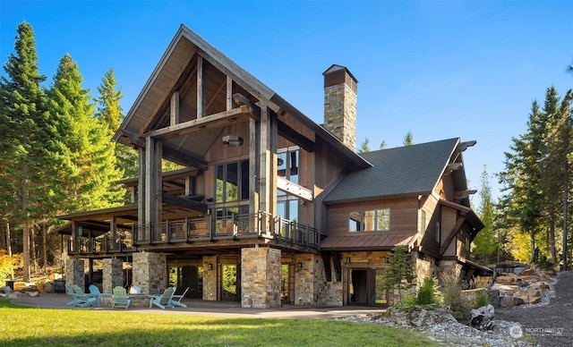 rear view of house featuring a shingled roof, stone siding, a lawn, a chimney, and a patio