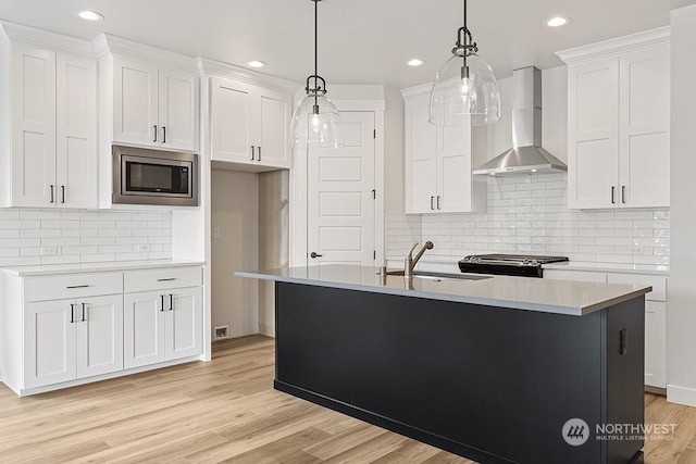 kitchen featuring stainless steel microwave, wall chimney exhaust hood, decorative light fixtures, a kitchen island with sink, and white cabinets