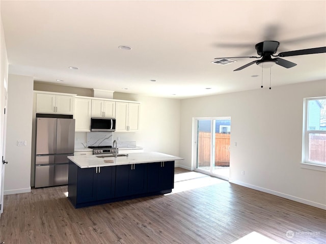 kitchen with backsplash, a kitchen island with sink, white cabinets, light wood-type flooring, and appliances with stainless steel finishes