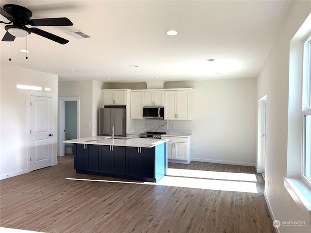 kitchen with ceiling fan, stainless steel appliances, an island with sink, white cabinets, and light wood-type flooring