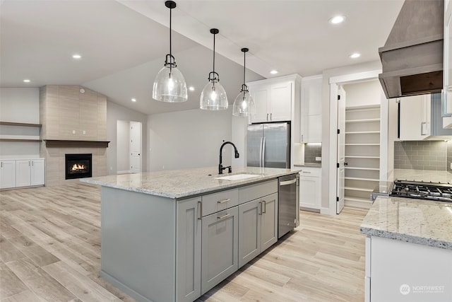 kitchen with decorative light fixtures, appliances with stainless steel finishes, a kitchen island with sink, a sink, and premium range hood