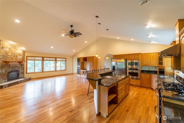 kitchen featuring appliances with stainless steel finishes, a stone fireplace, a kitchen island with sink, ceiling fan, and hanging light fixtures