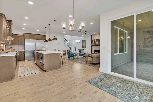 kitchen with ceiling fan, a stone fireplace, built in appliances, a kitchen island with sink, and light wood-type flooring