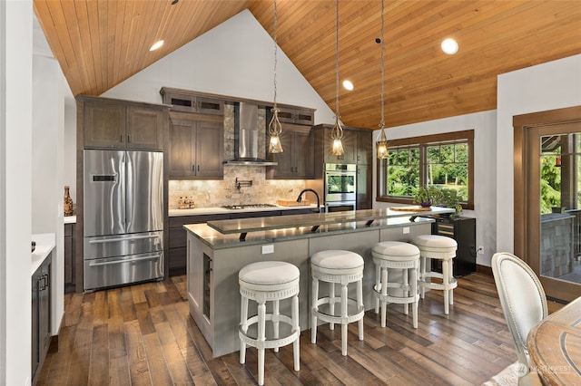 kitchen featuring wall chimney exhaust hood, hanging light fixtures, a center island with sink, and appliances with stainless steel finishes